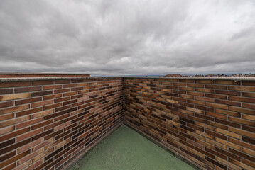 A brick wall of various shades of brown with a green cement floor on an attic terrace on a day with clouds heavy with water wanting to rain