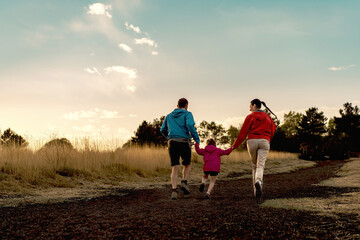 Naklejka na ściany i meble Sunset Run in the Mountains: A Mediterranean family, with mom, dad, and daughter, runs joyfully on a path, captured from behind in warm sunset light, wearing windbreakers.