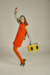 excited woman in orange dress dancing with yellow boombox and raised hand on grey, vintage vibes