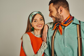 joyful woman in orange dress looking at camera near man in suspenders on grey, retro-inspired style