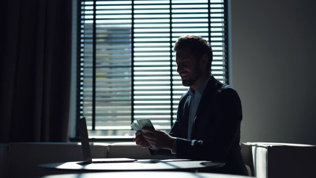 Successful business person sitting at work desk with wireless computer in dark stylish office and counting dollar bills. Cheerful man rejoicing money from profitable deal and kissing banknotes.