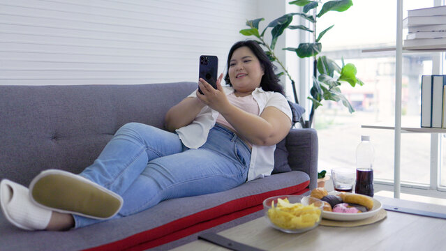 Cheerful Asian chubby woman smiling on the sofa, holding potato chips, taking selfie on smartphone.