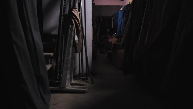 An abandoned warehouse with clothes. Clothes hang on hangers in covers. The camera is moving slowly through the rows. First person view. There are boxes on the floor. Dim light, scary is inside