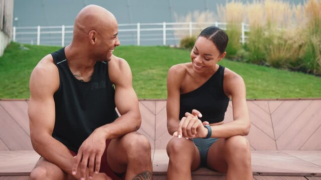 Pleased athletic african couple looking at wristwatch and talking after running near waterfront
