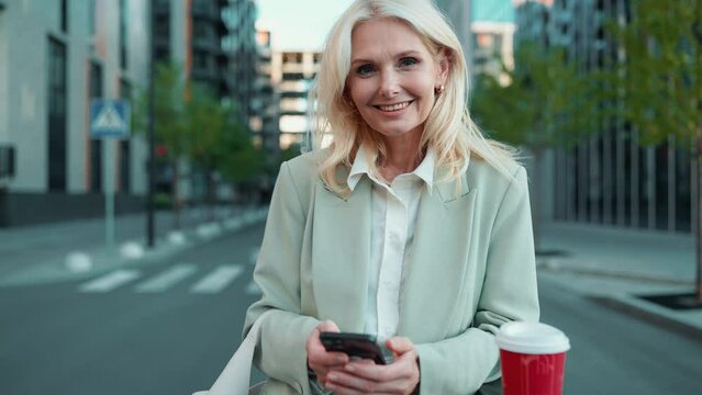 Cheerful blonde mature businesswoman in suit texting on cell phone and looking at camera with cup of coffee outdoors
