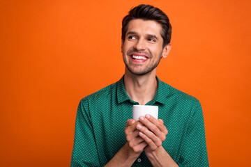 Photo of dreamy cheerful guy wear green shirt drinking cacao looking empty space isolated orange color background