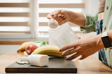 Woman in jeans and shirt with a measuring spoon in her hand puts portion of whey protein powder...