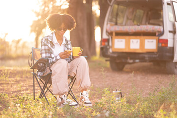 Young African black woman drinking a coffee and using her smartphone in the nature during a road trip with her camper van in autumn