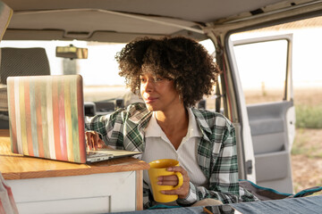 Young African black woman using her laptop inside her camper van during an autumn journey living a nomadic life