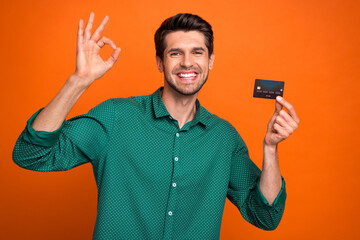 Photo of positive funky guy wear green shirt showing okey rising bank card isolated orange color background