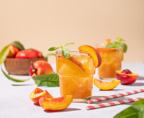 Two glasses with peach tea with ice and mint. Vegetarian and healthy homemade cold drink on a light background with fresh fruits and shadows.