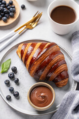 Fresh croissant with chocolate on a plate on a light background with a blueberries and cup of coffee close up. Delicious homemade breakfast concept.