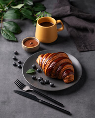 Freshly baked chocolate croissant with blueberries on a gray plate on a dark background with a yellow cup of black coffee. The concept of a delicious homemade breakfast in rustic style