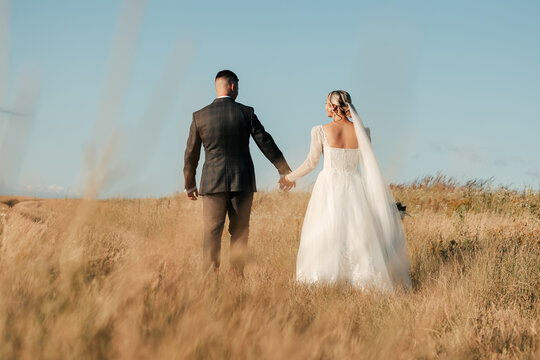 shot of a beautiful bride in a white dress and the groom holding her hand in the middle of a sunny day in the field, wedding photography, newlyweds walking together, love,common future