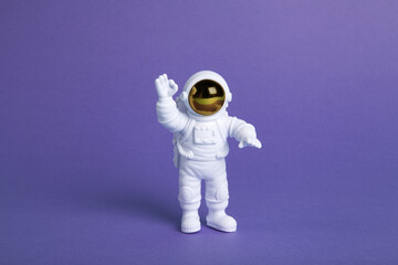 an astronaut figurine in a spacesuit exploring a plain purple background - obrazy, fototapety, plakaty