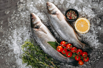 Two fresh seabass fish, which are located on ice particles on the kitchen table in one direction, next to which there is a black pot with pepper, sprigs of fragrant rosemary,