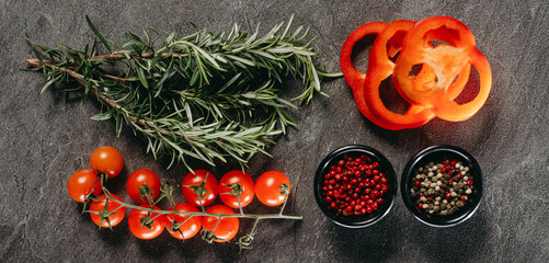 An exquisite arrangement of sprigs of fragrant rosemary, three rings of red pepper, a sprig of cherry tomatoes and two black cups with mixed pepper peas and red on a stone table