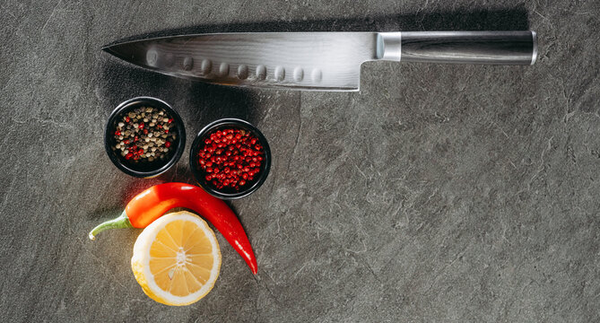 A large sharp kitchen knife with a black handle, next to two black cups with red pepper and mix, hot red pepper and a cut piece of lemon, which are on a kitchen stone table