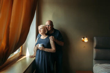 A happy elderly couple stand near the window, look