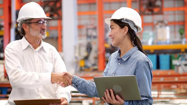 4k Video of happy senior male manager engineer wearing hardhat shaking hand with young beautiful programmer engineer holding a laptop in factory. colleague of different age, diversity teamwork concept
