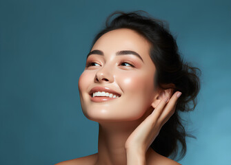Skin care. Woman with beauty face touching healthy facial skin portrait. Beautiful smiling asian girl model with natural makeup touching glowing hydrated skin on blue background closeup  - obrazy, fototapety, plakaty