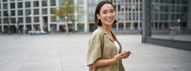 Portrait of asian woman standing on street, city square and holding mobile phone. Girl with smartphone walking outdoors