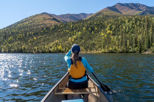 Woman paddling a canoe on a remote Yukon lake with beautiful scenery around her; Yukon, Canada