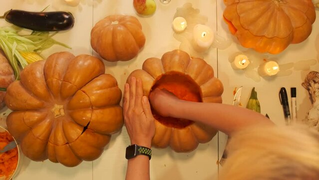 Top view of hands blonde girl who takes out the pulp from a pumpkin on a decorated table among corn and flickering candles in preparation for Halloween. Happy autumn holiday background.