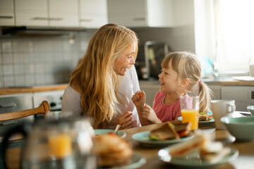 Young mother embracing her daughter while having breakfast together in the kitchen at home - obrazy, fototapety, plakaty
