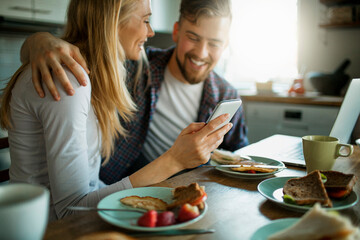 Young couple using a smartphone while having breakfast together in the morning in the kitchen at home - obrazy, fototapety, plakaty