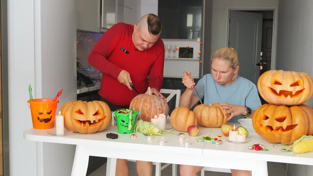 Young family in modern light kitchen intently carving jack-o'-lanterns from large pumpkins preparing to celebrate saints night holiday happy halloween festival background.