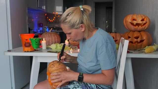 Side view young woman sitting in kitchen hold pumpkin in hands and drawing black felt-tip pen face preparation for carving Jack-o-lantern against background festive attributes Halloween holiday autumn