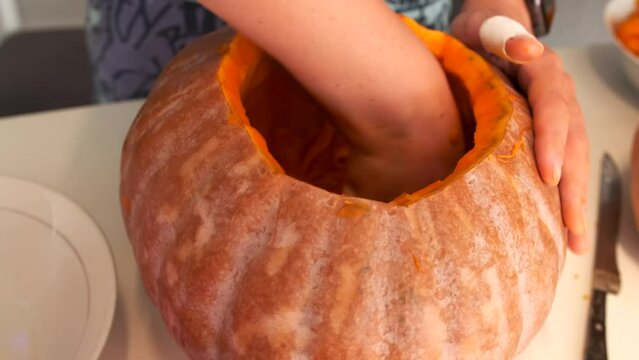 Close-up of female hands with a bandaged finger scraping out the core of the pulp and seeds from a large traditional pumpkin with a spoon for carving a Jack-o-lantern, preparing Halloween decorations.