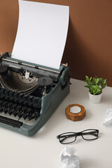 Vintage typewriter with candle, houseplant and eyeglasses on white table near brown wall