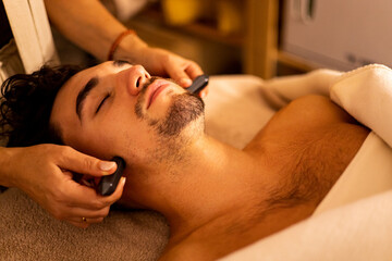 Obraz na płótnie Canvas A young adult with a beard is lying with his eyes closed on a stretcher in a beauty center undergoing a biotherapy session. The beautician places hematite stones on your face for blood stimulation.