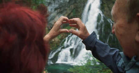 Older retired couple making heart with their hands while hiking to a waterfall in Austria. The concept of love to travel at any age. - obrazy, fototapety, plakaty