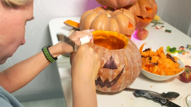 Young woman in kitchen table close-up carving face with knife large orange pumpkin along contour for Jack O lantern, preparing for celebration of Halloween decoration festival background tracking shot