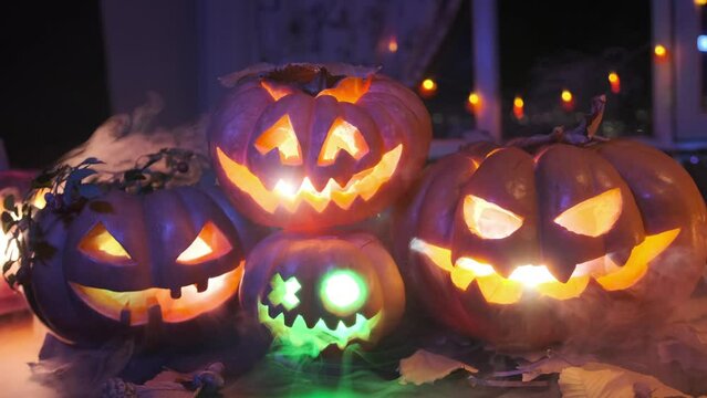 Tracking shot festive background for Halloween party invitation with four flashing lantern carved pumpkins in fog and in dry leaves among colorful garlands lying on the doorstep of house.