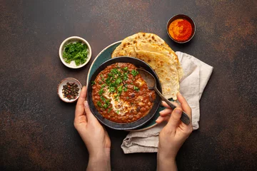 Zelfklevend Fotobehang Female hands holding a bowl and eating traditional Indian Punjabi dish Dal makhani with lentils and beans served with naan flat bread, fresh cilantro on brown concrete rustic table top view. © somegirl
