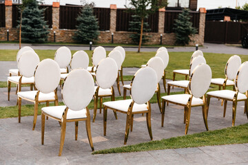Luxury white-golden chairs on wedding ceremony outdoors. Festive decorations. Empty rows armchairs for guests. Outgoing wedding ceremony in the open air. Decor Studio.