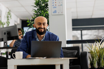 Smiling arab businessman working on laptop in business office while sitting in couch. Start up...