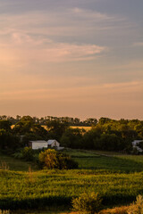 Obraz premium Green meadows with a rural white house and a small barn, against a backdrop of blue sky and chaotic orange clouds.
