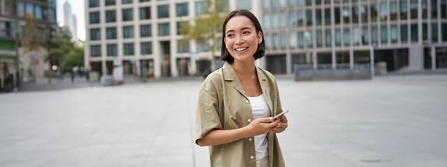 Portrait of asian woman standing on street, city square and holding mobile phone. Girl with...