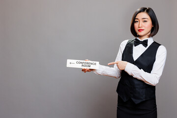 Asian woman receptionist holding conference room directional tabletop signage and pointing with finger portrait. Hotel employee in uniform showing boardroom steel sign and looking at camera