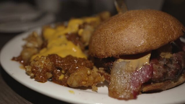 This mouth watering, panning video features a huge bacon burger with a side of chili cheese tots on a white plate at a restaurant.