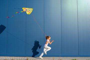 Joyful young European blonde girl in a white T-shirt and white sweatpants with pink heart-shaped glasses runs with a kite in her hand against a blue wall outside - 653990115