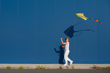 Joyful young European blond girl in a white T-shirt and white sweatpants with pink heart-shaped glasses flies a kite against a blue wall outside - 653990139