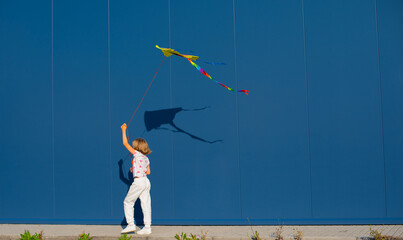 Joyful young European blond girl in a white T-shirt and white sweatpants with pink heart-shaped glasses flies a kite against a blue wall outside - 653990156