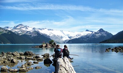 A young couple, sitting on a log overlooking a small rocky island, enjoys the tranquility of a snowy glacier and the quiet blue of Lake Garibaldi, British Columbia, Canada.