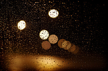 View through the rainy window at night with water drops and blurry city lights.